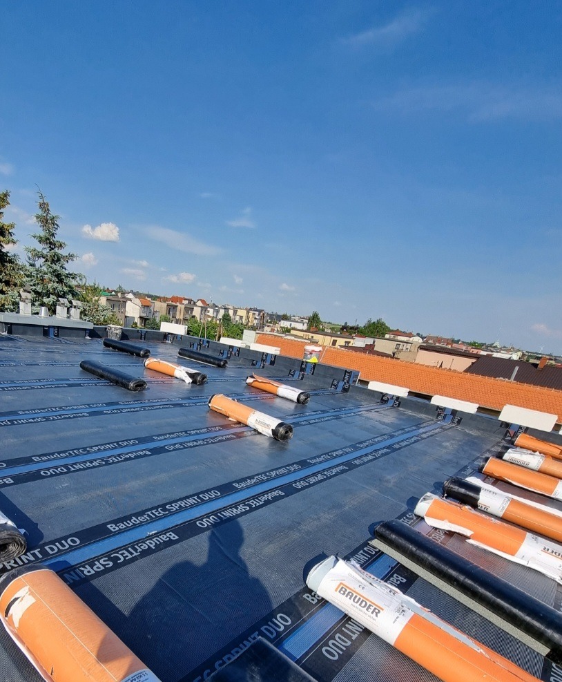 Rozwinięte rolki papy termozgrzewalnej BauderTEC Sprint DUO na płaskim dachu w trakcie prac dekarskich, widok z góry na tle zabudowy miejskiej i błękitnego nieba.