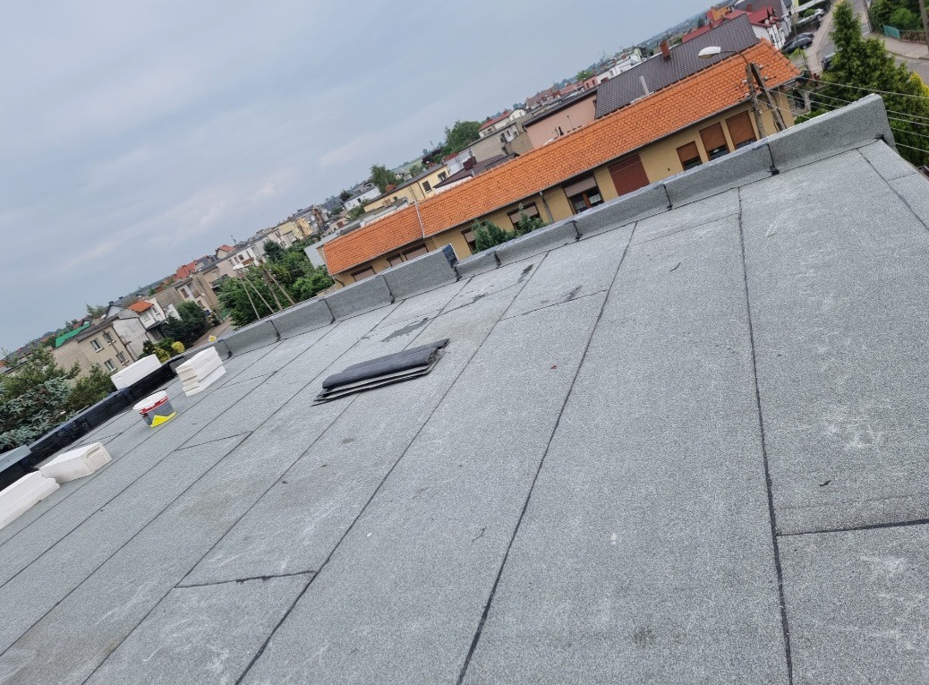 Płaski dach pokryty papą termozgrzewalną z kominkiem wentylacyjnym, widok na zabudowę miejską w tle, częściowo przygotowany do prac wykończeniowych, z widocznym pojemnikiem z masą bitumiczną...