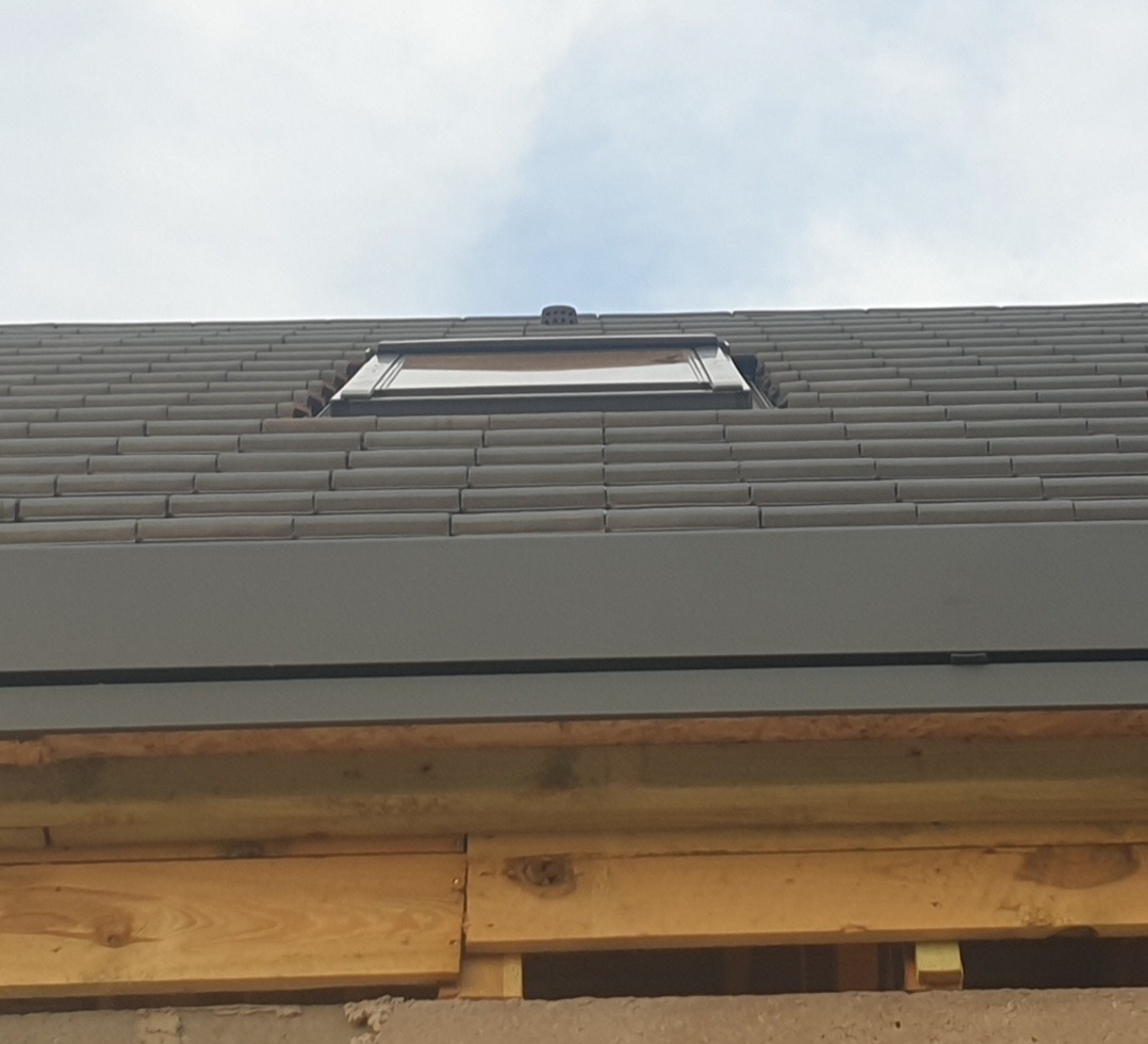 Szare dachówki ceramiczne z oknem dachowym i obróbką blacharską, widoczne drewniane elementy konstrukcji dachu.