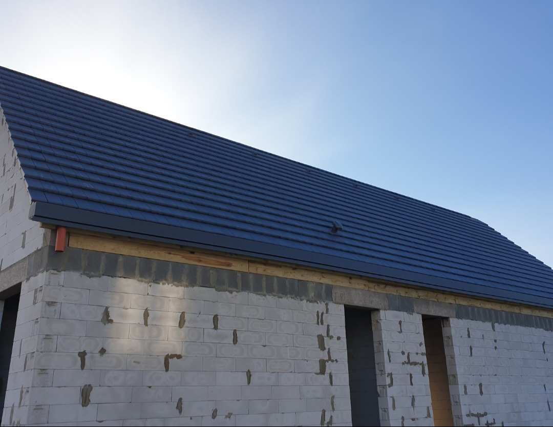 Nowy, grafitowy dach z dachówki ceramicznej na surowym budynku z białego bloczka, widoczna pomarańczowa rura spustowa.