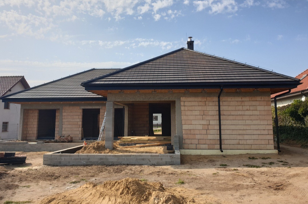 Nowy dom w Gostyniu w trakcie budowy, z ciemną dachówką i czarnymi rynnami, widoczne elementy konstrukcyjne.