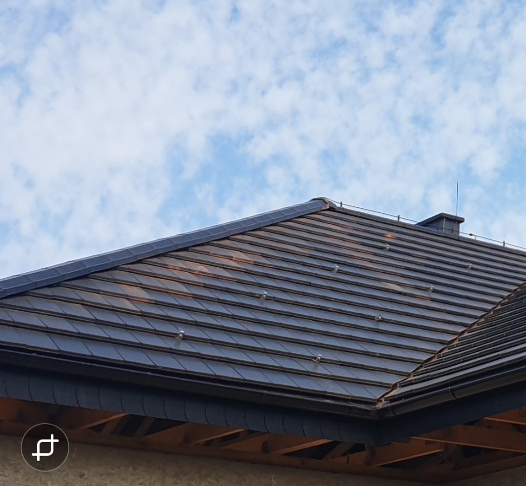 Ciemnobrązowy dach ceramiczny z widocznymi klamrami mocującymi, kominem i fragmentem nieba z chmurami.