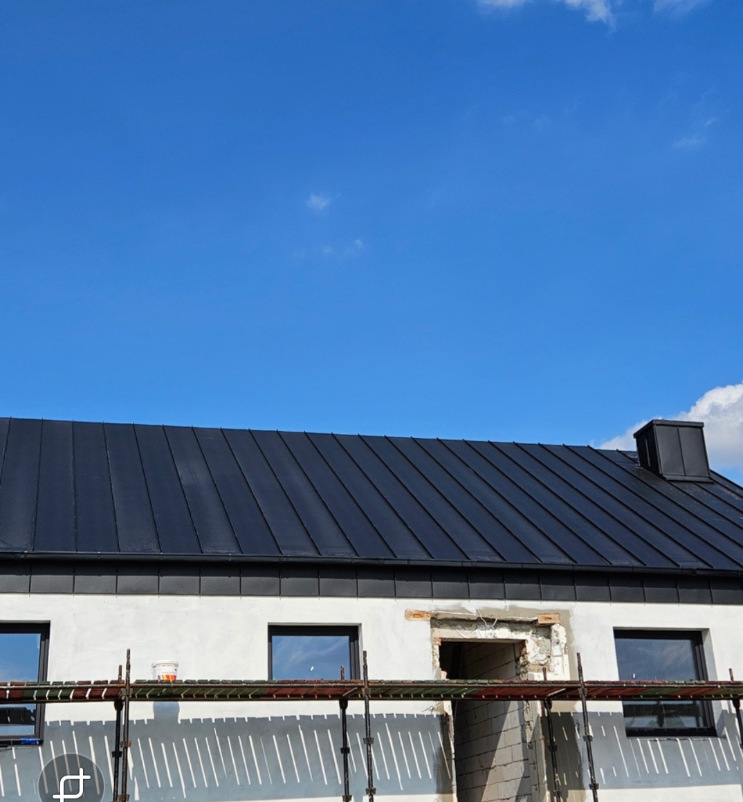 Dom jednorodzinny z nowym, czarnym dachem z blachy na rąbek stojący, widoczny fragment rusztowania i otwór okienny w trakcie prac wykończeniowych.