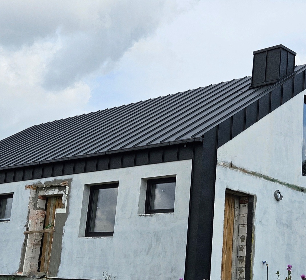 Nowoczesny dom z czarnym dachem z blachy na rąbek stojący i białymi ścianami, widoczne niedokończone wykończenia okien i drzwi.