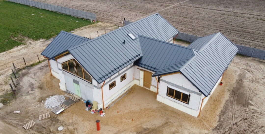 Dom w trakcie budowy z nowym, szarym dachem z blachy na rąbek stojący, widok z drona, z widocznymi rynnami i oknami z drewnianą stolarką, teren wokół w trakcie przygotowania.