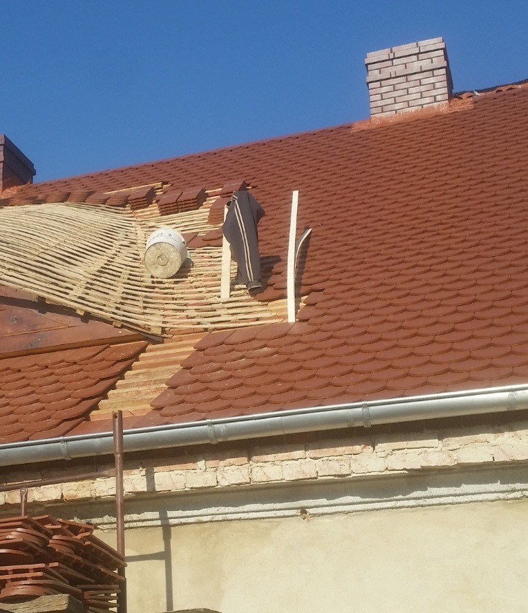 Częściowo zdjęty dach z brązową dachówką, widoczne łaty, wiadro i robocze ubranie na dachu, komin z cegły w tle