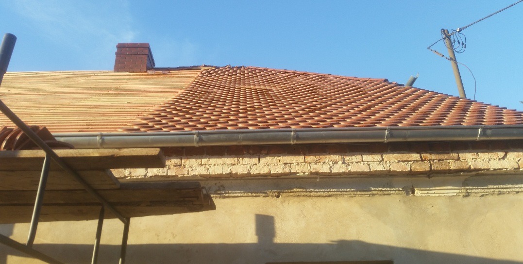 Częściowo położony dach z czerwonej dachówki karpiówki, widoczny komin i fragment rusztowania, na połowie dachu widoczna konstrukcja drewniana pod dachówką.