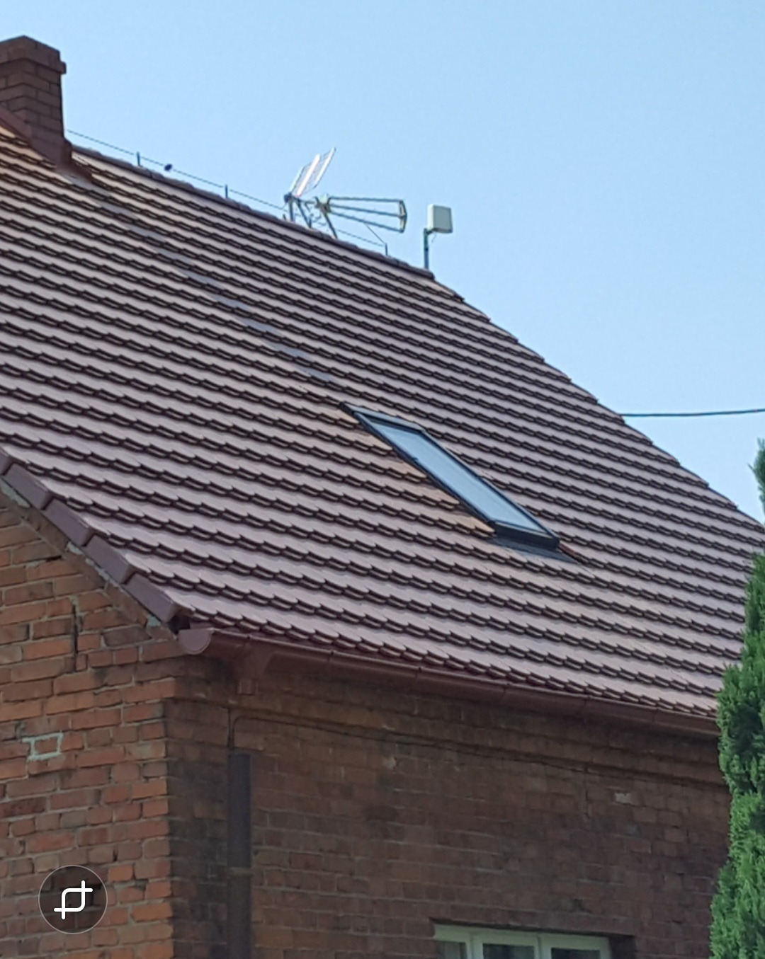 Czerwony dach z dachówki ceramicznej na ceglanym domu z oknem dachowym i anteną telewizyjną na szczycie.