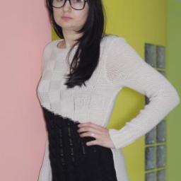 Z.P.U.H. Toma - Kobieta w okularach prezentuje dzianinową sukienkę z kontrastującym, ukośnym połączeniem kremowej góry i czarnego dołu z warkoczowym splotem, stojąc na tle ściany w pastelowych kolorach.