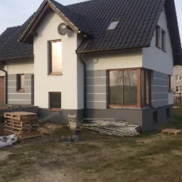 Nowoczesny dom jednorodzinny z szarą elewacją, widoczne detale wykończeniowe i materiały budowlane. Dom z brązowymi oknami i czarnym dachem.