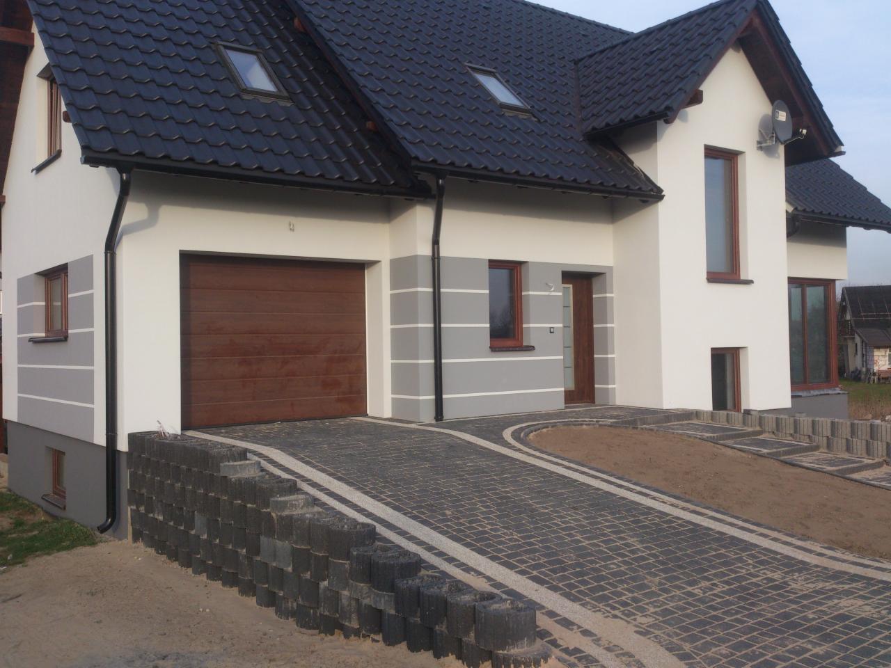 Dwukondygnacyjny dom z brązową bramą garażową i ciemnym dachem. Elewacja w odcieniach szarości i bieli. Podjazd z kostki brukowej.