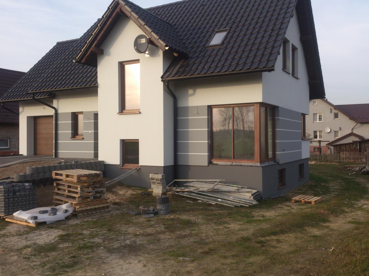 Nowoczesny dom jednorodzinny z szarą elewacją, widoczne detale wykończeniowe i materiały budowlane. Dom z brązowymi oknami i czarnym dachem.