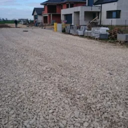 Przygotowany podkład z kruszywa pod budowę drogi do nowo powstających domów jednorodzinnych, widoczne palety z kostką brukową i elementy betonowe obrzeży.