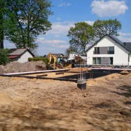 Wykop pod fundamenty domu jednorodzinnego z widocznym zbrojeniem, izolacją przeciwwilgociową i żółtą koparką w tle, teren przygotowany do wylewania betonu.