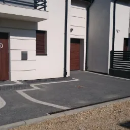 Nowoczesny podjazd z szarej kostki brukowej z białymi akcentami, prowadzący do drzwi wejściowych i bramy przesuwnej, widok na dwukondygnacyjny dom z balkonem.