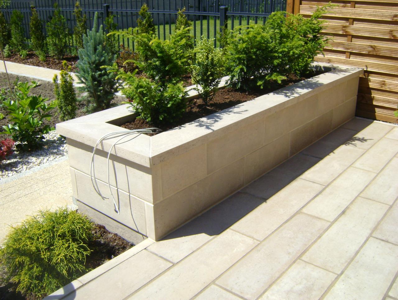 Beżowa donica z kostki brukowej, wypełniona zielenią, na tle jasnej kostki brukowej. Widoczne ogrodzenie i drewniana ściana w tle. Kabel zwisający z donicy.