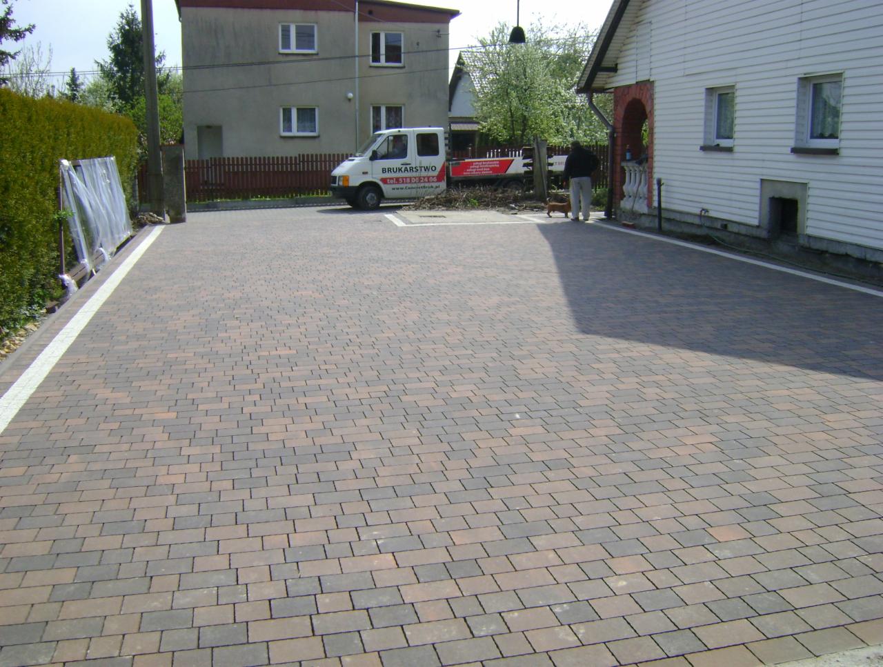 Ułożona kostka brukowa w odcieniach brązu i szarości tworzy wzór na podjeździe. Biały pas obrzeża oddziela kostkę od zieleni i budynku. Na drugim planie budynki i zaparkowany bus.