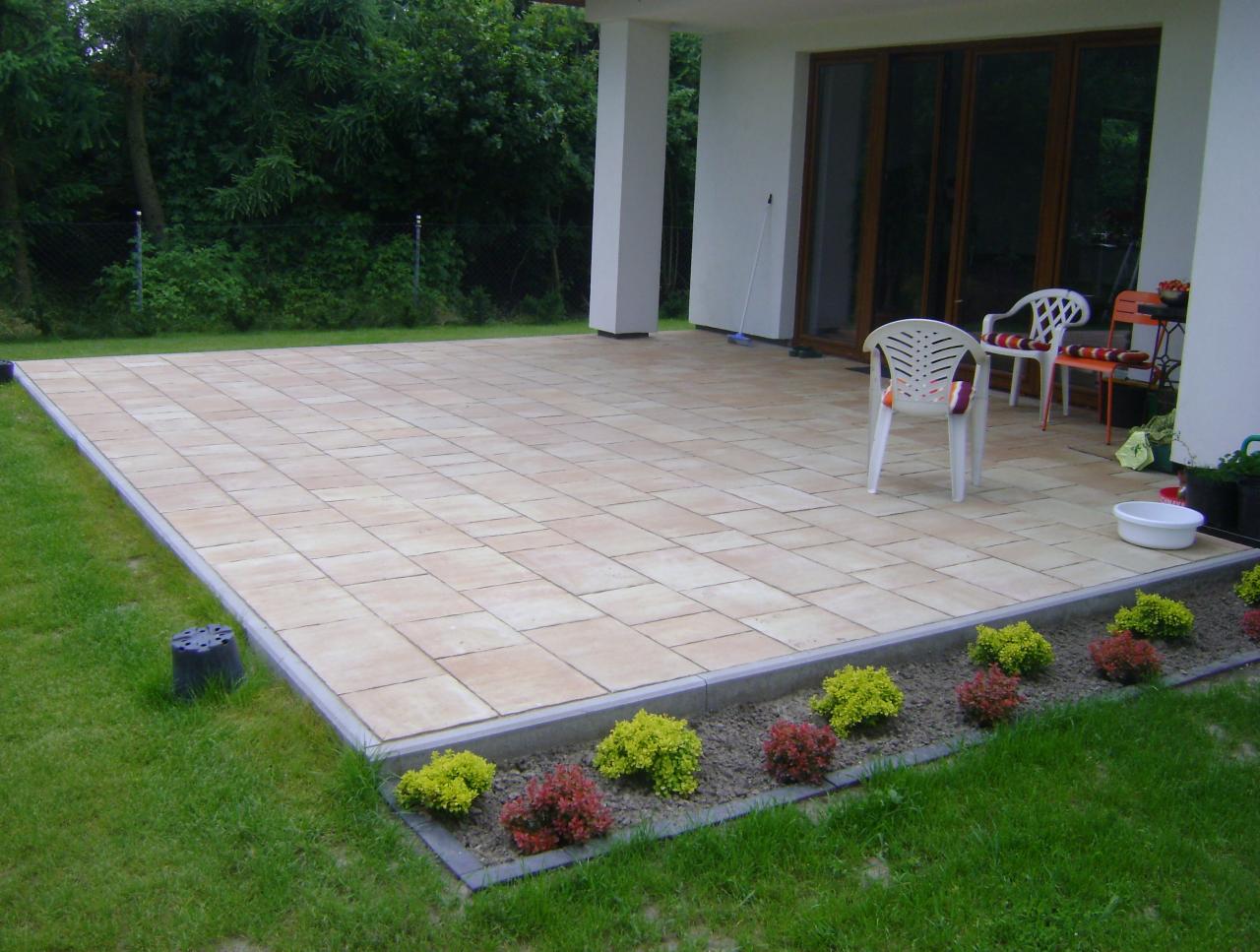 Jasna, prostokątna kostka brukowa tworząca taras przy domu. Widoczne krzesła, rośliny ozdobne i trawnik. Ujęcie z niskiej perspektywy podkreśla regularność wzoru.