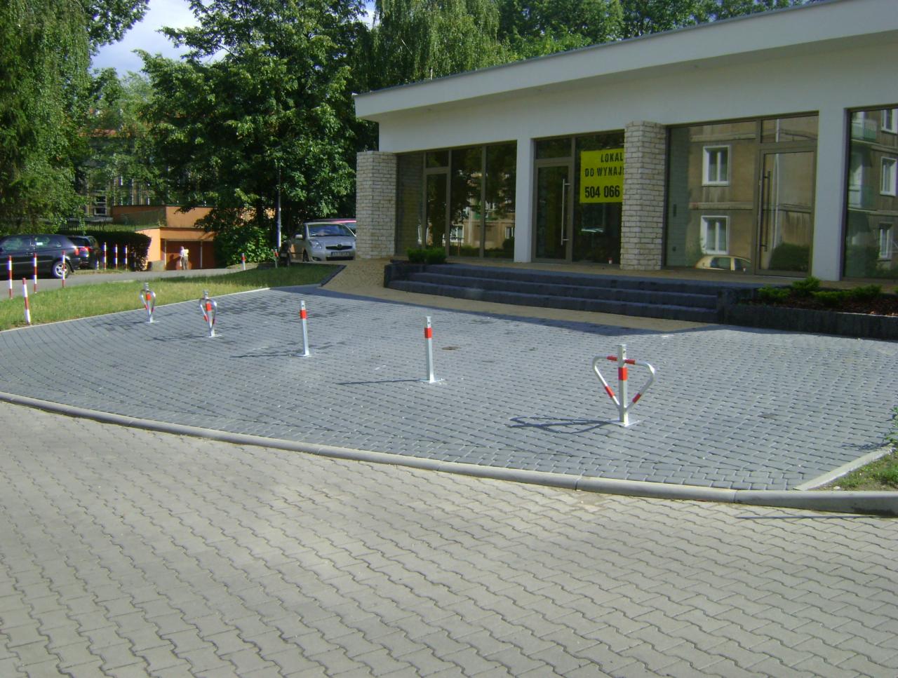 Szara kostka brukowa na parkingu przed budynkiem z przeszkleniami, zabezpieczona słupkami i blokadą parkingową. Widoczne schody wejściowe i zieleń w tle.