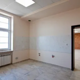 Pusta kawiarenka w budynku biurowym z oknem, grzejnikiem, częściowo wyłożona kafelkami, z widocznymi przyłączami wodnymi i otwartymi drzwiami do kolejnego pomieszczenia z ceglaną ścianą.