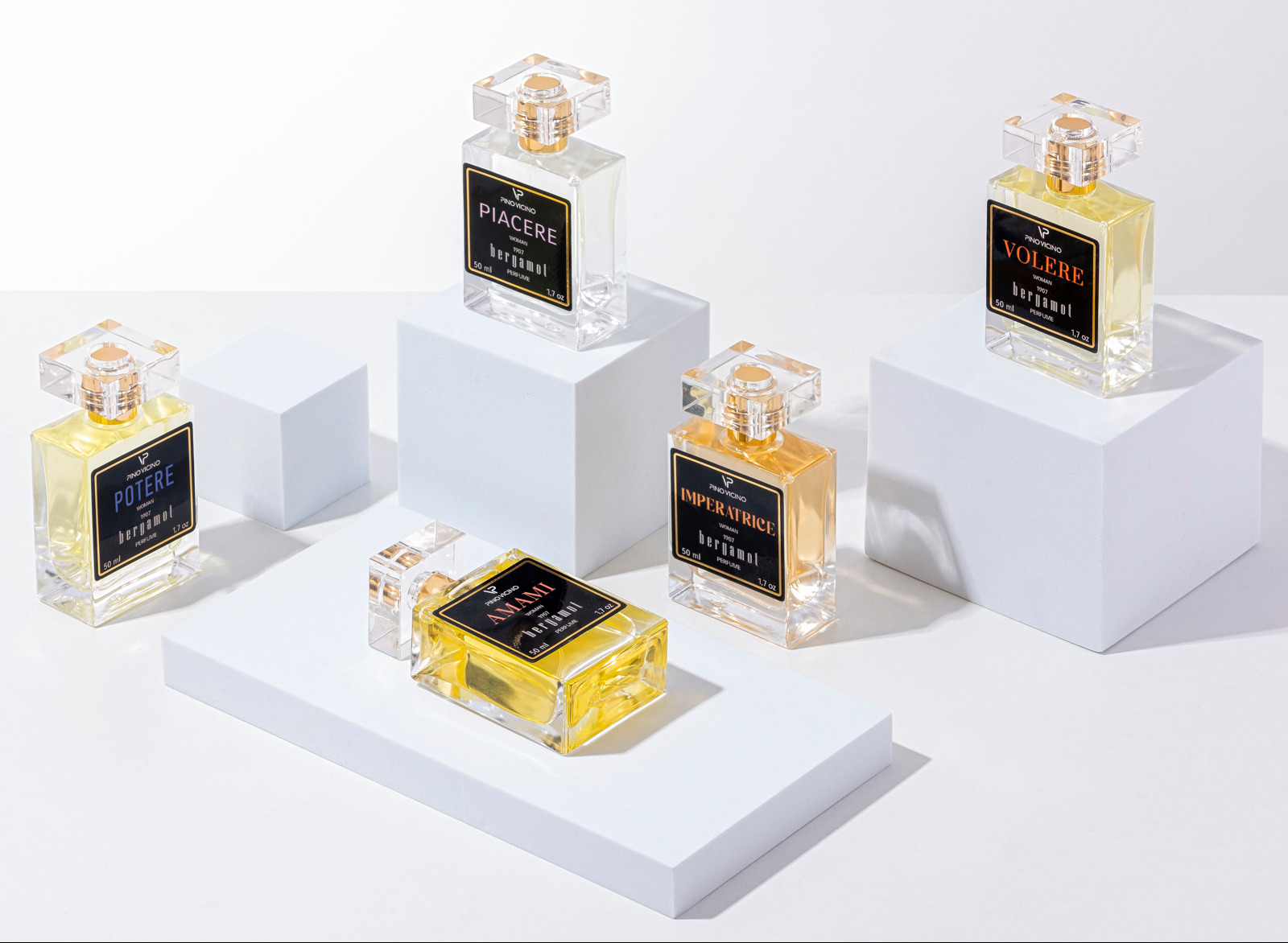 Kompozycja pięciu flakonów perfum Pino Vicino o różnych zapachach (Potere, Piacere, Amami, Imperatrice, Volere) na białych platformach i kostkach, fotografia reklamowa na jasnym tle.