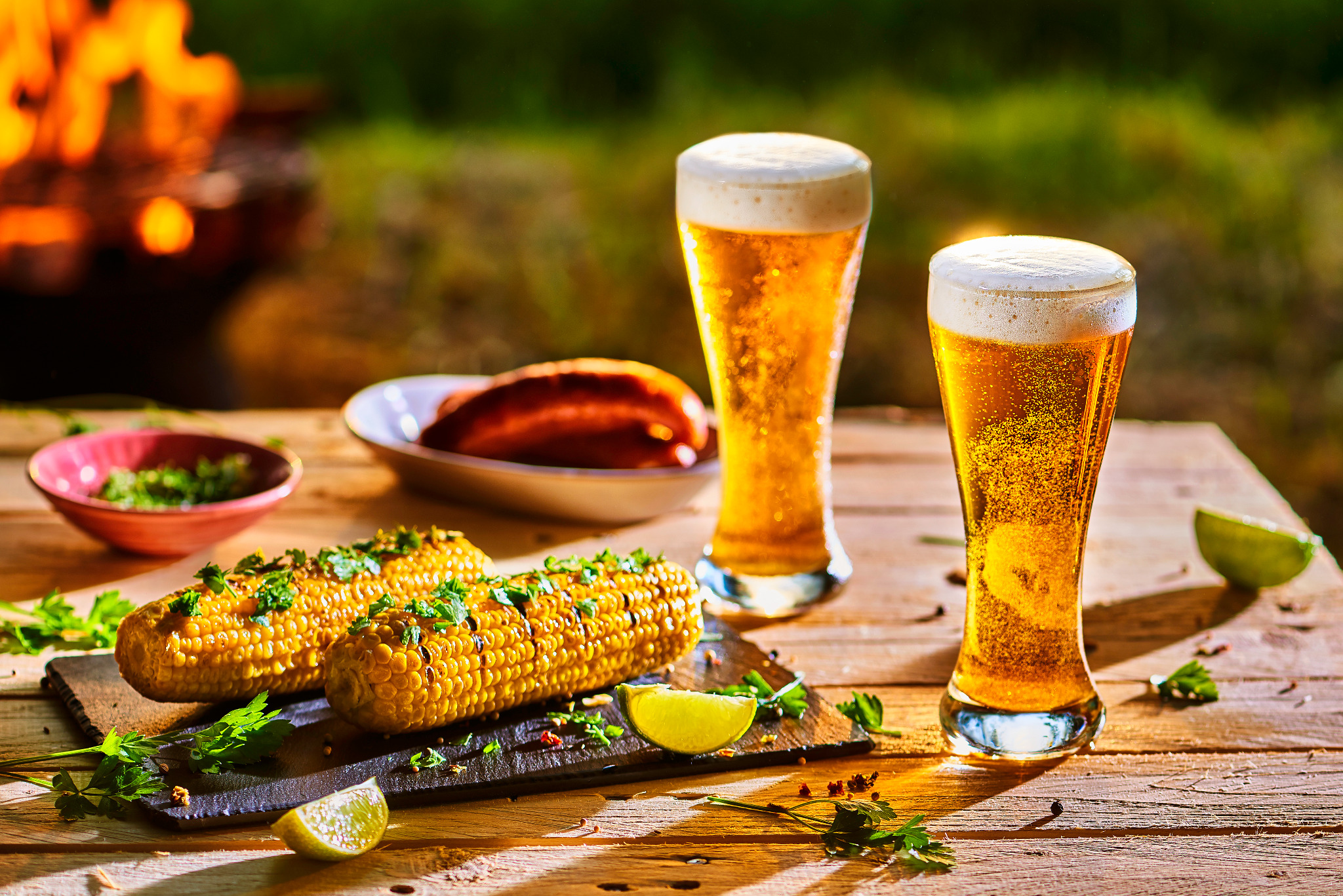 Dwa kufle jasnego piwa z pianką i grillowana kukurydza z ziołami i limonką na drewnianym stole, w tle ognisko.