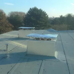 Płaski dach pokryty papą z dwoma kominami wentylacyjnymi zakończonymi daszkami, w tle budynek mieszkalny i drzewa.