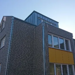 Nowoczesny budynek z szarej cegły z metalowym dachem w kolorze grafitowym i żółtymi panelami balkonowymi, widoczne rynny w kolorze srebrnym.