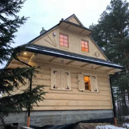 Nowy dom z bali w stylu góralskim, z drewnianymi okiennicami i podświetlonymi oknami, częściowo wykończony, z widoczną fundamentową ścianą i elementami instalacji na zewnątrz, otoczony drzewami...