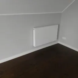 Biały, płaski grzejnik panelowy zamontowany na szarej ścianie pod skosem poddasza, widoczne gniazdka elektryczne i ciemna podłoga.