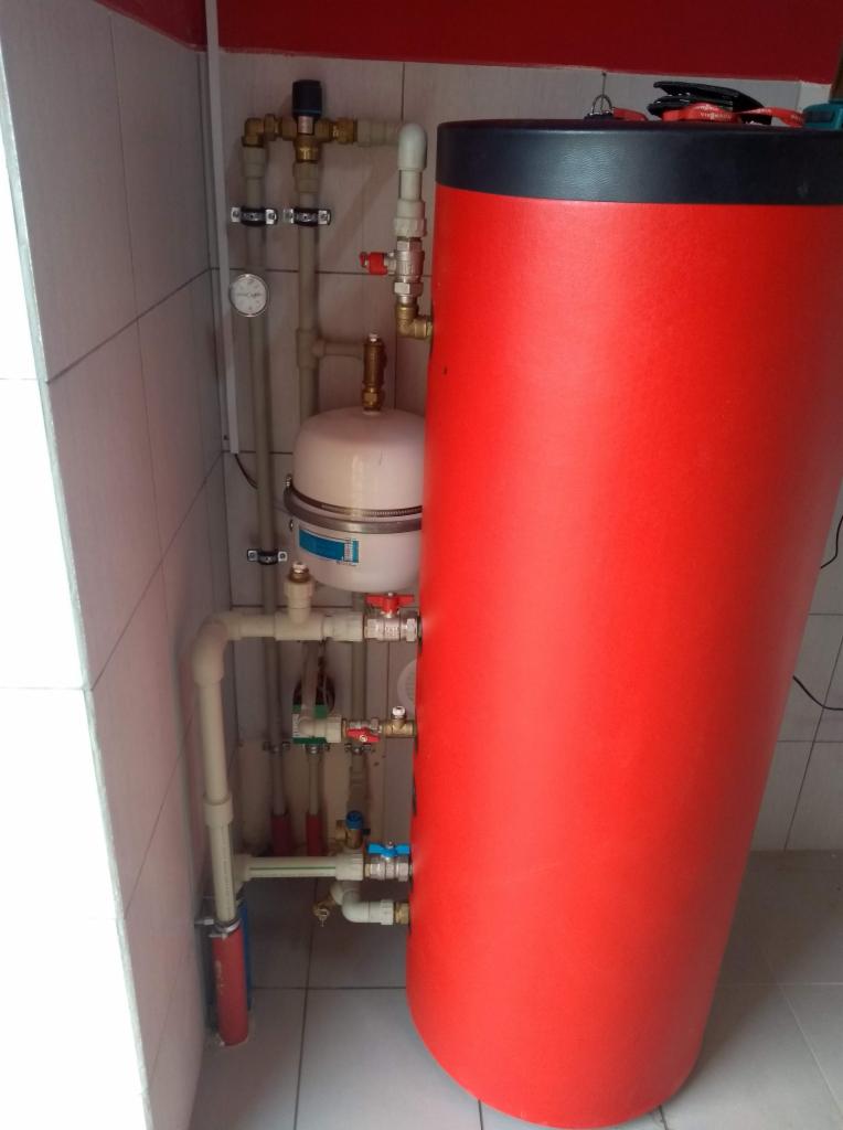 Czerwony, cylindryczny zasobnik ciepłej wody z instalacją hydrauliczną i manometrem, zamontowany w rogu pomieszczenia z białymi płytkami.