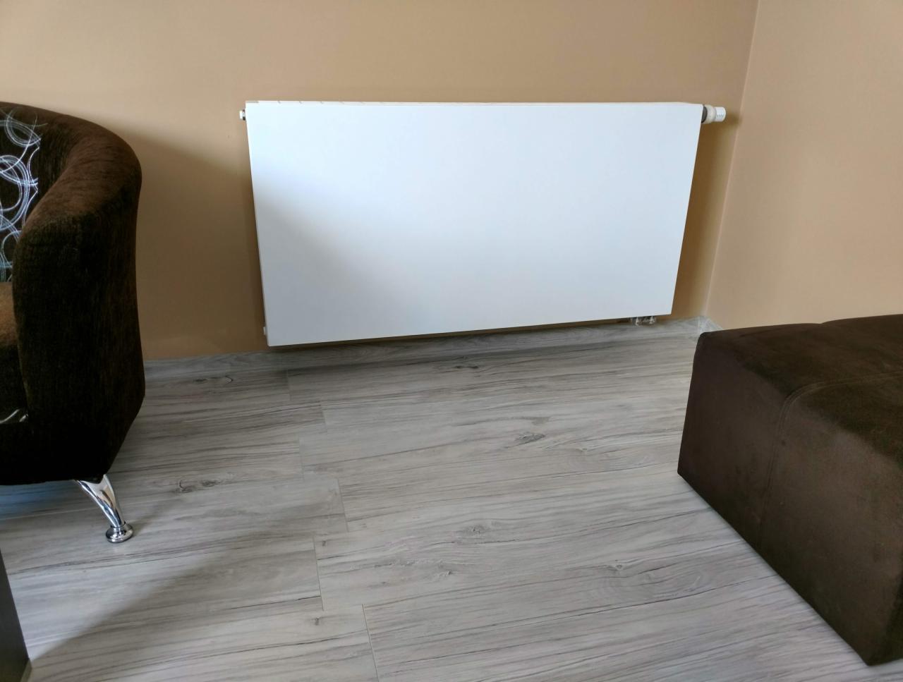 Biały, płaski grzejnik panelowy zamontowany na beżowej ścianie, widoczny fragment brązowego fotela z chromowaną nogą i pufa na szarych panelach podłogowych.