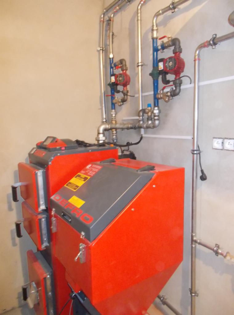 Czerwony piec na paliwo stałe marki DEFRO z szarym panelem sterowania i metalowymi rurami instalacji grzewczej na tle szarej ściany.