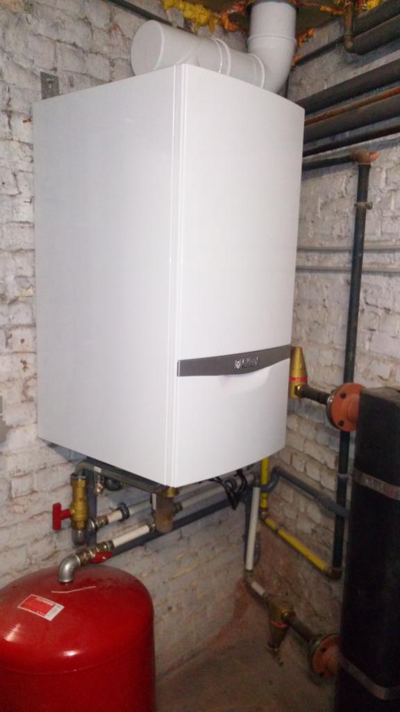 Biały, wiszący piec gazowy marki Vaillant z podłączonymi rurami na tle ściany z cegły, widoczny również czerwony zbiornik wyrównawczy.