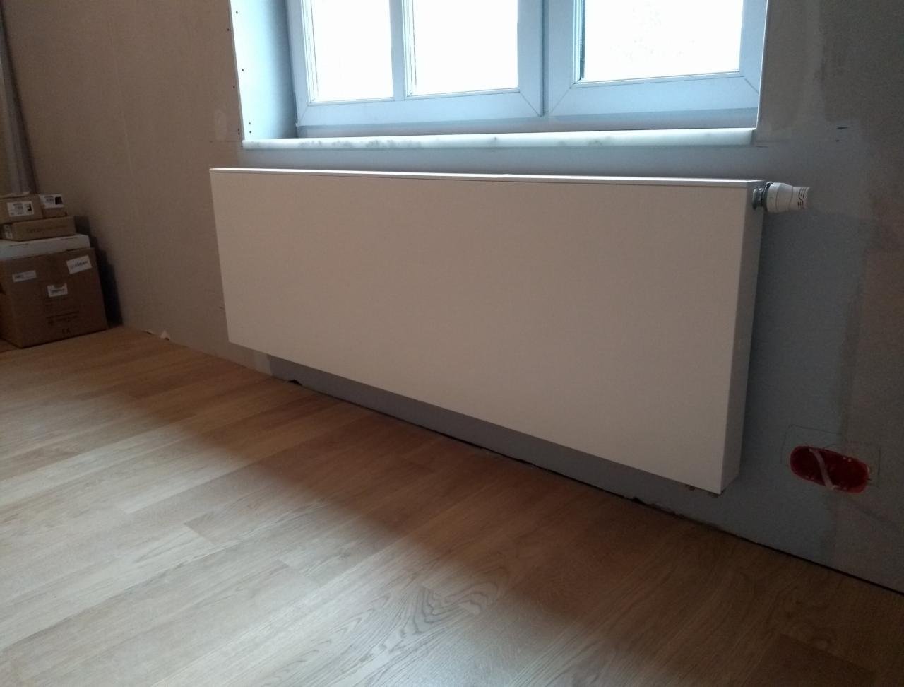 Biały, płaski grzejnik panelowy zamontowany pod oknem na tle surowej ściany w trakcie wykończenia wnętrza, widoczna drewniana podłoga i kartony w rogu pokoju.