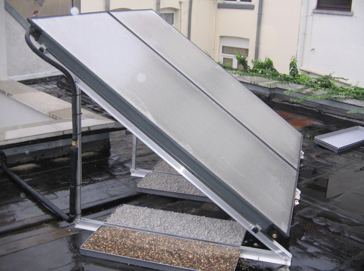 Kolektor słoneczny na dachu budynku, z widocznymi rurami przyłączeniowymi i zasobnikami z kamieniami pod konstrukcją wsporczą.