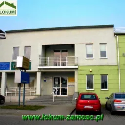 Dwupiętrowy budynek biurowy z kilkoma firmami, w tym agencją nieruchomości, z zaparkowanymi samochodami przed wejściem i logo 'LOKUM' w górnym rogu.