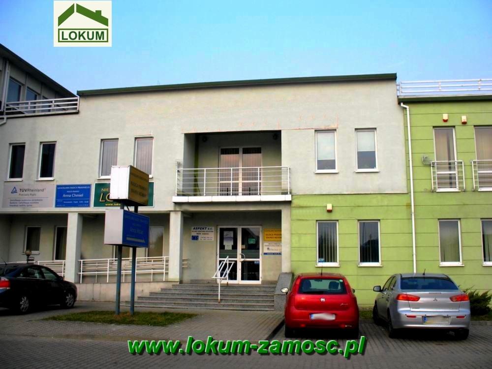 Dwupiętrowy budynek biurowy z kilkoma firmami, w tym agencją nieruchomości, z zaparkowanymi samochodami przed wejściem i logo 'LOKUM' w górnym rogu.