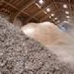 Góra jasnego, sypkiego materiału, prawdopodobnie biomasy, składowana wewnątrz hali z drewnianą konstrukcją dachu i oświetleniem przemysłowym.