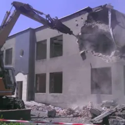 Żółta koparka z młotem pneumatycznym rozbija szary, dwukondygnacyjny budynek. Gruz i pył unoszą się w powietrzu. Widoczne niezabezpieczone rusztowania.