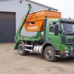 Zielona ciężarówka Volvo z pomarańczowym kontenerem na gruz, zaparkowana przed szarą halą z zamkniętą bramą. Widoczny numer kontaktowy na kontenerze.