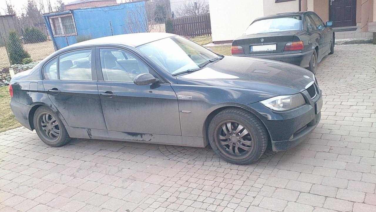 Dwa ciemne, brudne samochody BMW zaparkowane na kostce brukowej. Jeden z nich, z widocznymi zabrudzeniami na karoserii, stoi bliżej, drugi w tle.