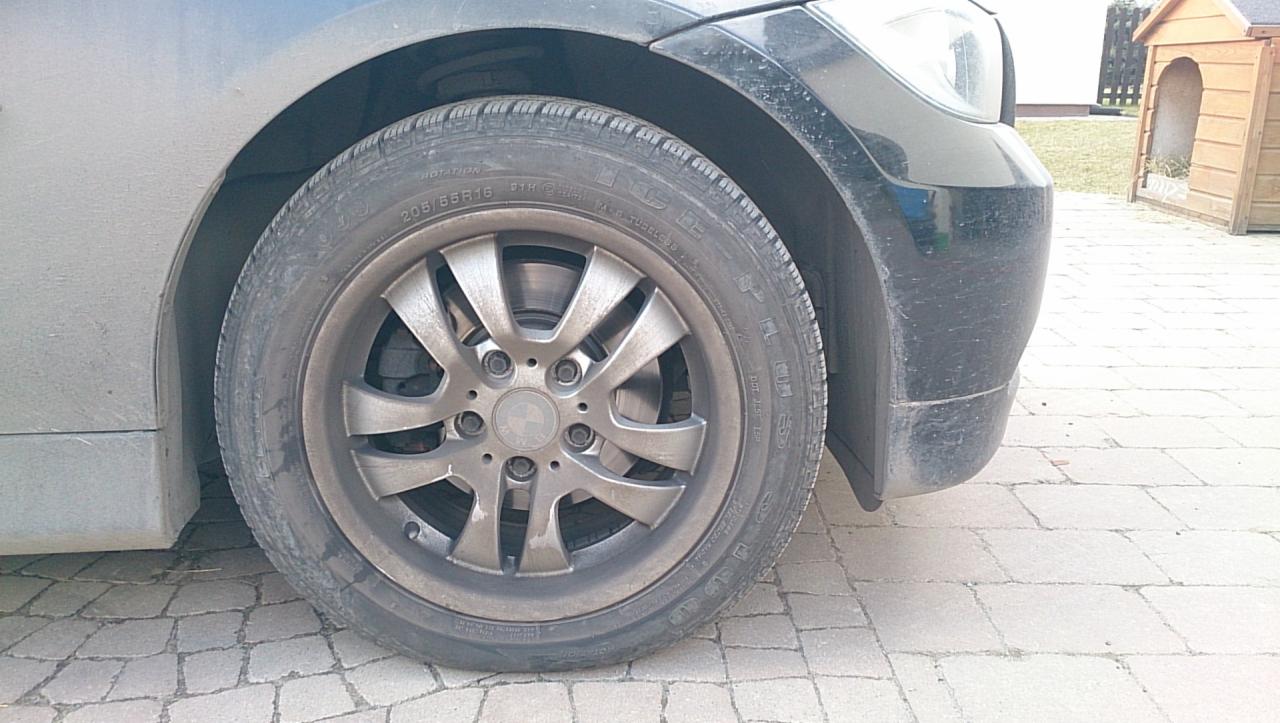 Błotniste koło BMW na kostce brukowej, widoczne zabrudzenia na oponie i feldze, w tle fragment domu i buda dla psa. Auto wymaga umycia.