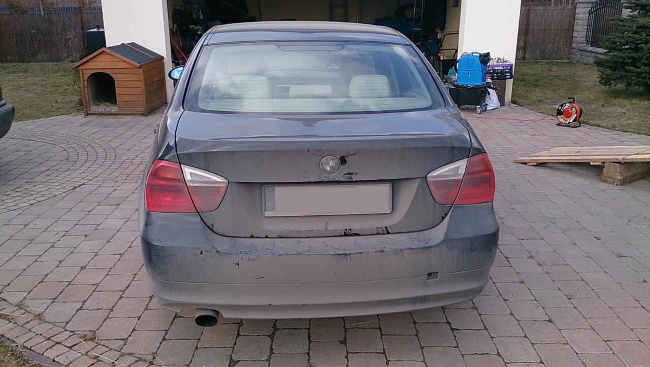 Widok z tyłu brudnego, ciemnego sedana BMW na kostce brukowej. W tle garaż, buda dla psa i zwinięty przedłużacz elektryczny.