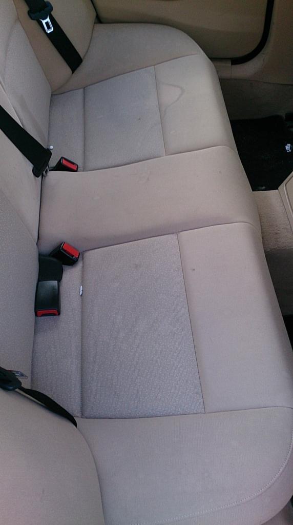 Beżowa, materiałowa tylna kanapa samochodowa z widocznymi plamami i zabrudzeniami na siedzisku i oparciu, pasy bezpieczeństwa.