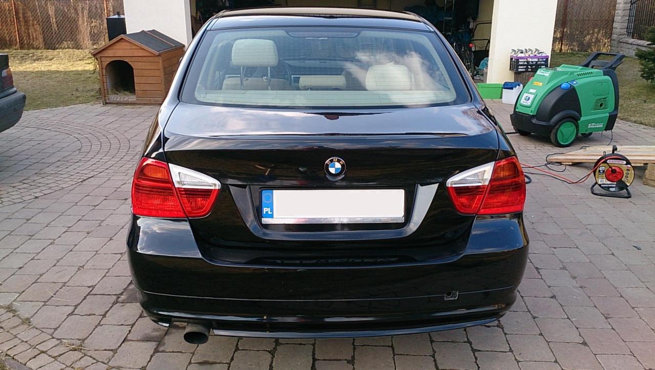 Tył czarnego BMW na kostce brukowej z zieloną myjką ciśnieniową i budą dla psa w tle. Tablica rejestracyjna z polską flagą. Widok z tyłu na czysty lakier.