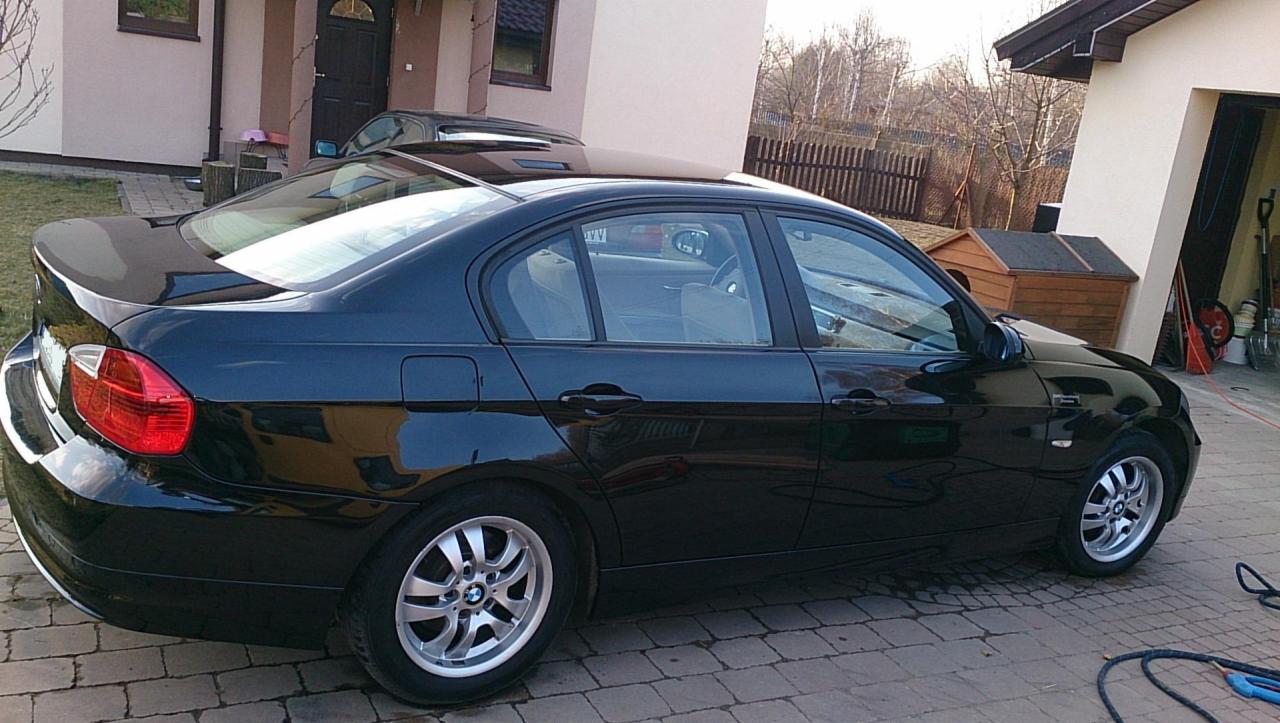 Błyszczący, czarny sedan BMW po umyciu na kostce brukowej. Widoczne refleksy światła na karoserii, garaż w tle. Auto zaparkowane na posesji.