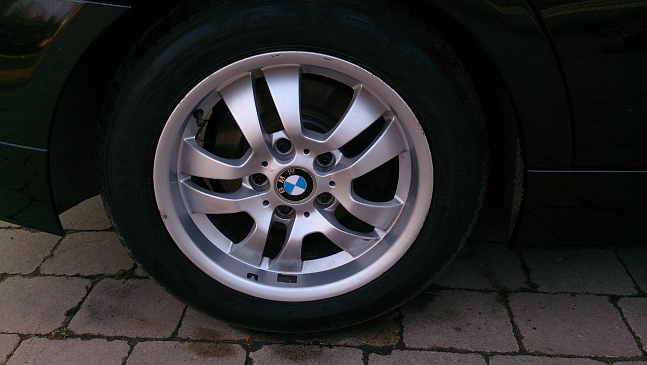 Aluminiowa felga BMW z oponą na tle kostki brukowej. Widoczne detale bieżnika opony i wzór felgi. Auto w kolorze czarnym.