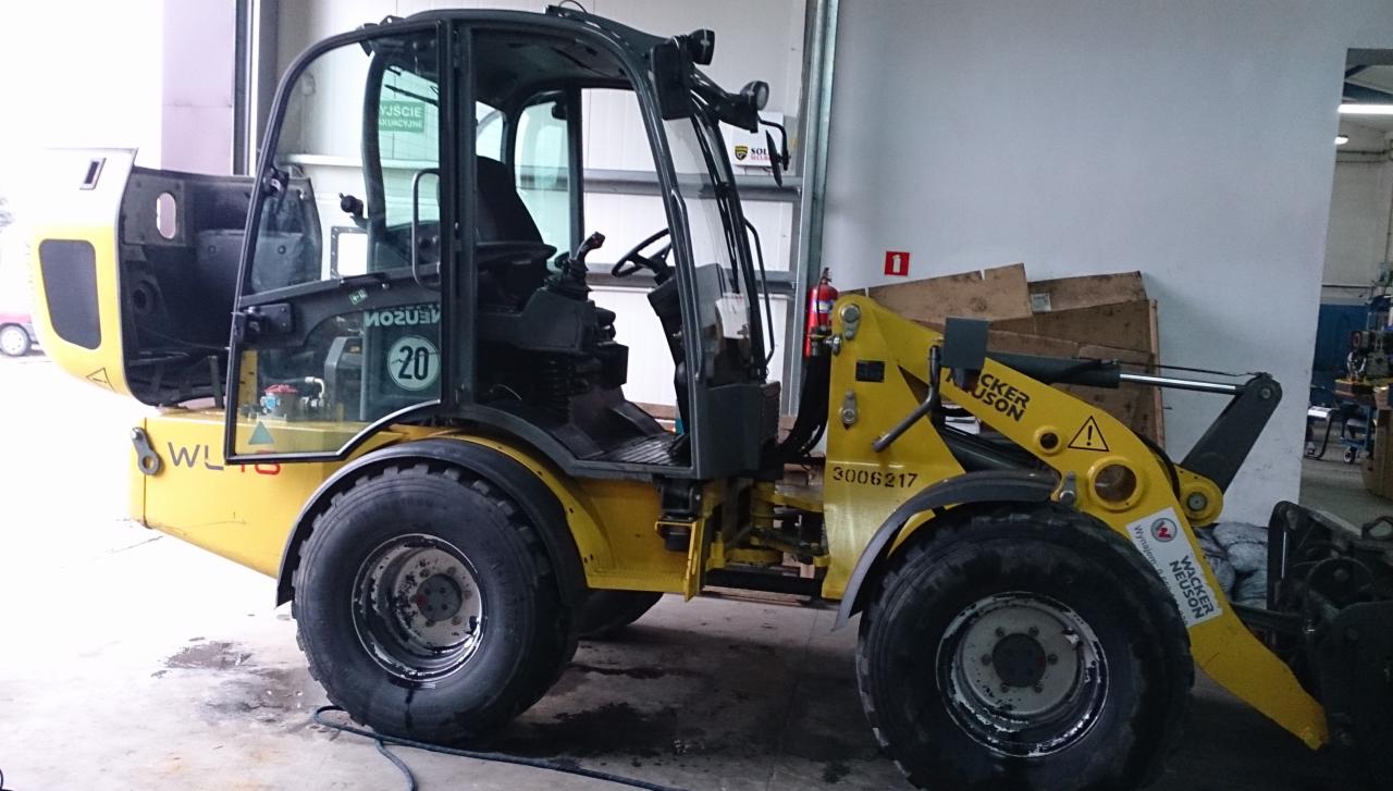 Żółta ładowarka Wacker Neuson WL18 z otwartą kabiną, widoczny numer 20 na szybie, zaparkowana w hali. Otwarta kabina i widoczne detale konstrukcyjne.