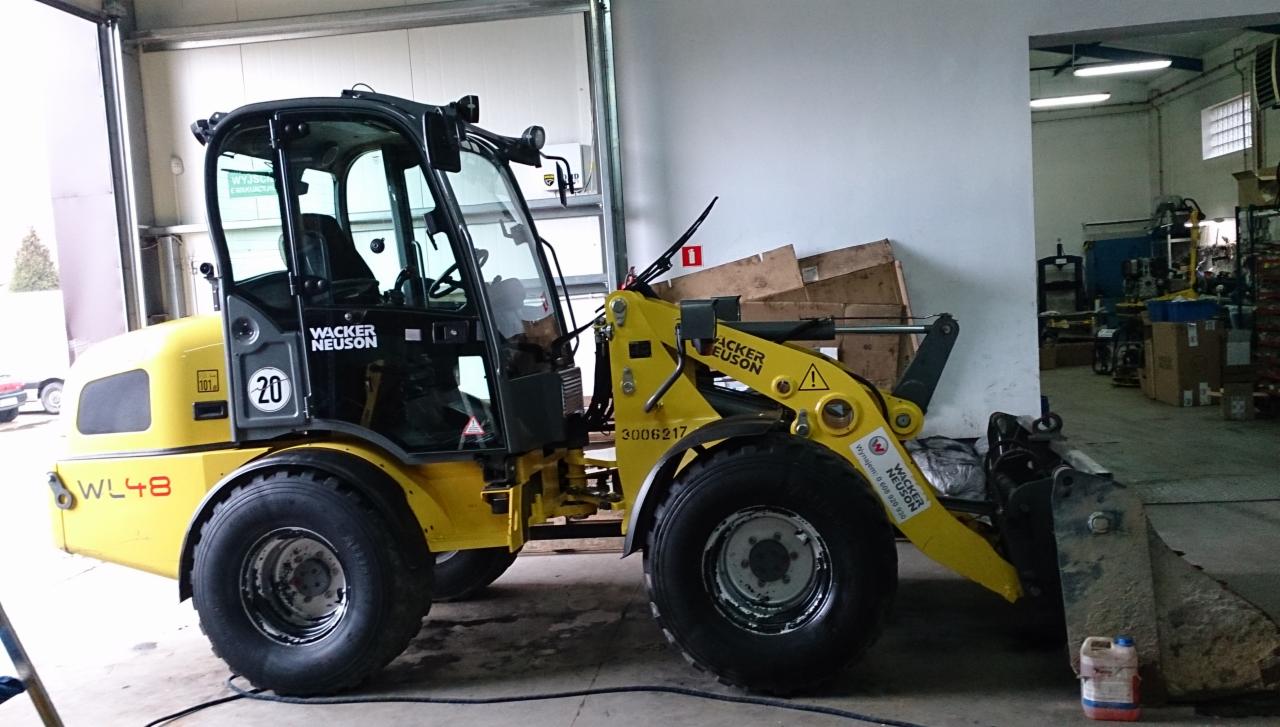 Żółta ładowarka Wacker Neuson WL48 w garażu, widok z boku. Maszyna budowlana z łyżką, gotowa do pracy. W tle skrzynie i wyposażenie warsztatu.